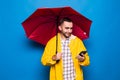 Young handsome bearded man in yellow raincoat with red umbrella use mobile phone isolated over blue background Royalty Free Stock Photo