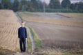 Young handsome blond child boy standing alone on ground road among golden grassy wheat fields on blurred foggy green trees and