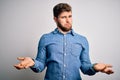 Young handsome blond man with beard and blue eyes wearing casual denim shirt clueless and confused with open arms, no idea concept Royalty Free Stock Photo