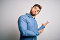 Young handsome blond man with beard and blue eyes wearing casual denim shirt Inviting to enter smiling natural with open hand Royalty Free Stock Photo