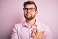 Young handsome blond man with beard and blue eyes wearing pink shirt and glasses smiling with hands on chest with closed eyes and Royalty Free Stock Photo