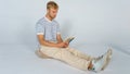 Young handsome blond man in casual T-shirt and beige pants on a white background reading a book sitting on a cushion on the floor