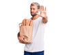 Young handsome blond man holding paper bag with bread with open hand doing stop sign with serious and confident expression, Royalty Free Stock Photo