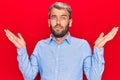 Young handsome blond man wearing casual shirt standing over isolated red background clueless and confused with open arms, no idea Royalty Free Stock Photo