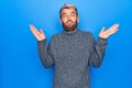 Young handsome blond man wearing casual sweater standing over isolated blue background clueless and confused with open arms, no Royalty Free Stock Photo