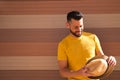 A young, handsome, blue-eyed, bearded man with a yellow T-shirt holds a hat in his hands. In the background a wall in shades of Royalty Free Stock Photo