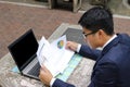 Young handsome business man is analyzing financial charts and documents against laptop in the public outdoors park. Royalty Free Stock Photo