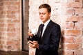 Young handsome businessman in black suit is drinking coffee and surfing in the phone near brick wall Royalty Free Stock Photo