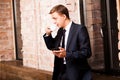 Young handsome businessman in black suit is drinking coffee and surfing in the phone near brick wall Royalty Free Stock Photo