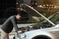 Young handsome businessman standing near open front car bumper, checking engine , looks upset, unexpectable vehicle trouble