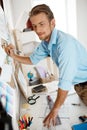 Young handsome businessman standing at table and writing on the paper pinned to corkboard. Office background.