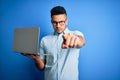 Young handsome businessman wearing glasses working using laptop over blue background pointing with finger to the camera and to Royalty Free Stock Photo