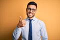 Young handsome businessman wearing tie and glasses standing over yellow background doing happy thumbs up gesture with hand Royalty Free Stock Photo