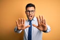 Young handsome businessman wearing tie and glasses standing over yellow background doing stop gesture with hands palms, angry and Royalty Free Stock Photo