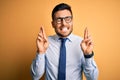 Young handsome businessman wearing tie and glasses standing over yellow background gesturing finger crossed smiling with hope and