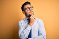 Young handsome businessman wearing tie and glasses standing over yellow background with hand on chin thinking about question, Royalty Free Stock Photo