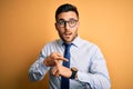 Young handsome businessman wearing tie and glasses standing over yellow background In hurry pointing to watch time, impatience, Royalty Free Stock Photo