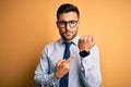 Young handsome businessman wearing tie and glasses standing over yellow background In hurry pointing to watch time, impatience, Royalty Free Stock Photo