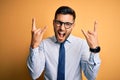 Young handsome businessman wearing tie and glasses standing over yellow background shouting with crazy expression doing rock Royalty Free Stock Photo