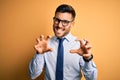 Young handsome businessman wearing tie and glasses standing over yellow background smiling funny doing claw gesture as cat,