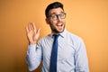 Young handsome businessman wearing tie and glasses standing over yellow background Waiving saying hello happy and smiling, Royalty Free Stock Photo