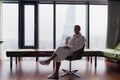 Young handsome carefree man wearing bathrobe near modern full length window enjoying a cup of coffee while looking Royalty Free Stock Photo