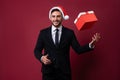 Young handsome caucasian guy in business suit and Santa hats stands on red background in studio and smilie throws up red gift box