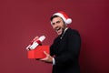 Young handsome caucasian guy in business suit and Santa hats stands on red background in studio and teeth smilie Holding red gift