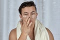 Young handsome caucasian man applying cream under the eyes, towel on shoulders. Caring face, metrosexual daily routine in the bath Royalty Free Stock Photo