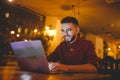 A young handsome Caucasian man with beard and toothy smile in a red checkered shirt is working behind a gray laptop sitting at a w Royalty Free Stock Photo
