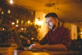 A young handsome Caucasian man with beard and toothy smile in a red checkered shirt is working behind a gray laptop sitting at a w Royalty Free Stock Photo