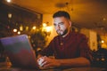 A young handsome Caucasian man with beard and toothy smile in a red checkered shirt is working behind a gray laptop Royalty Free Stock Photo