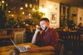 Young handsome Caucasian man with beard and toothy smile in red shirt works behind laptop, hands on keyboard sitting at wooden tab Royalty Free Stock Photo