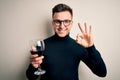 Young handsome caucasian man drinking an alcoholic glass of red wine over isolated background doing ok sign with fingers,