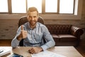 Young handsome cheerful businessman sit on table and pose in his own office. He hold big thumb up and smile. Nice happy