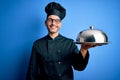 Young handsome chef man wearing uniform and hat holding waiter tray with dome with a happy face standing and smiling with a