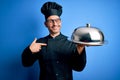 Young handsome chef man wearing uniform and hat holding waiter tray with dome with surprise face pointing finger to himself