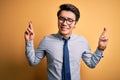 Young handsome chinese businessman wearing glasses and tie over yellow background gesturing finger crossed smiling with hope and