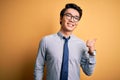 Young handsome chinese businessman wearing glasses and tie over yellow background smiling with happy face looking and pointing to Royalty Free Stock Photo