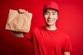 Young handsome chinese delivery man holding takeaway paper bag with food with a happy face standing and smiling with a confident Royalty Free Stock Photo