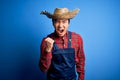 Young handsome chinese farmer man wearing apron and straw hat over blue background angry and mad raising fist frustrated and Royalty Free Stock Photo