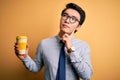 Young handsome chinese man drinking glass of take away coffee over yellow background serious face thinking about question, very