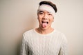 Young handsome chinese man injured for accident wearing bandage and strips on head sticking tongue out happy with funny expression