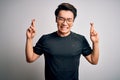 Young handsome chinese man wearing black t-shirt and glasses over white background gesturing finger crossed smiling with hope and Royalty Free Stock Photo