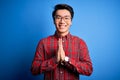 Young handsome chinese man wearing casual shirt and glasses over blue background praying with hands together asking for Royalty Free Stock Photo