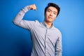 Young handsome chinese man wearing casual shirt standing over isolated blue background Strong person showing arm muscle, confident Royalty Free Stock Photo