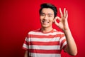 Young handsome chinese man wearing casual striped t-shirt standing over red background smiling positive doing ok sign with hand Royalty Free Stock Photo