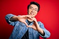 Young handsome chinese man wearing denim jacket and glasses over red background smiling in love doing heart symbol shape with Royalty Free Stock Photo