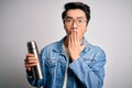 Young handsome chinese tourist man wearing glasses holding thermo with water cover mouth with hand shocked with shame for mistake,