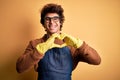 Young handsome cleaner man wearing apron and gloves over isolated yellow background smiling in love showing heart symbol and shape